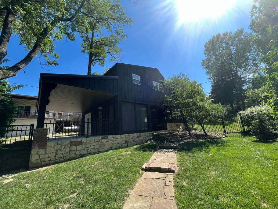 A renovated dark-colored barn turned vacation home with unique architectural features, nestled among green trees under a bright, sunny sky.