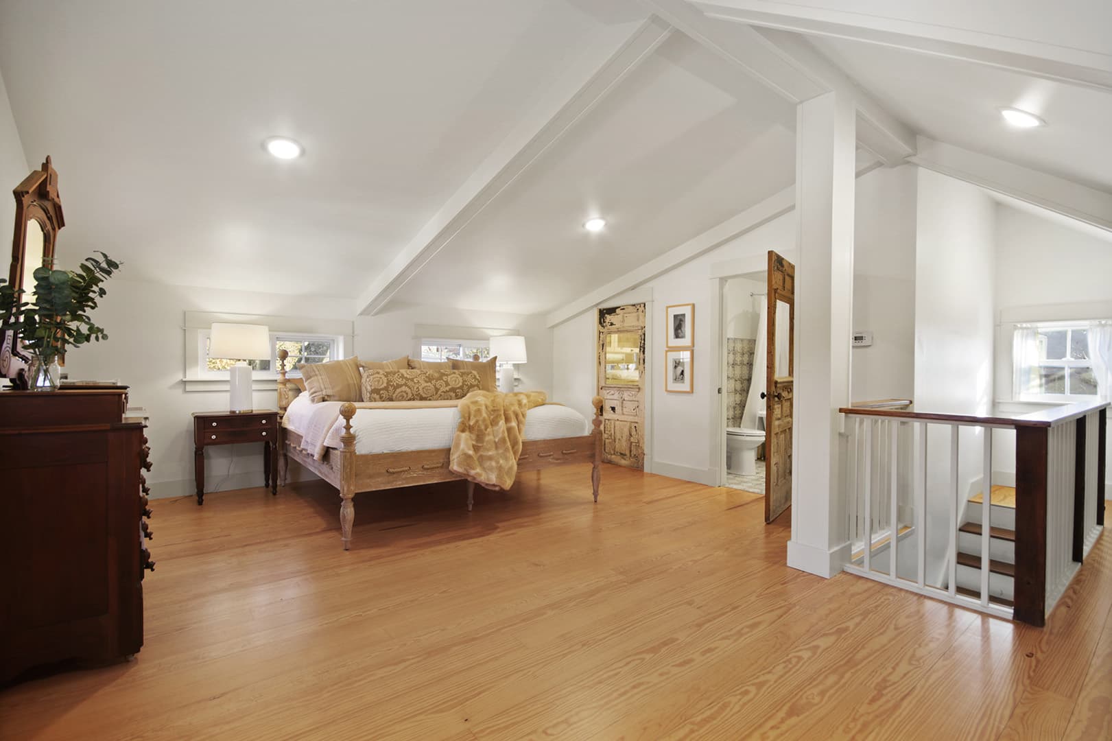A bright, spacious attic bedroom with a vaulted ceiling, light wood floors, a large bed, and a partial view of a bathroom.