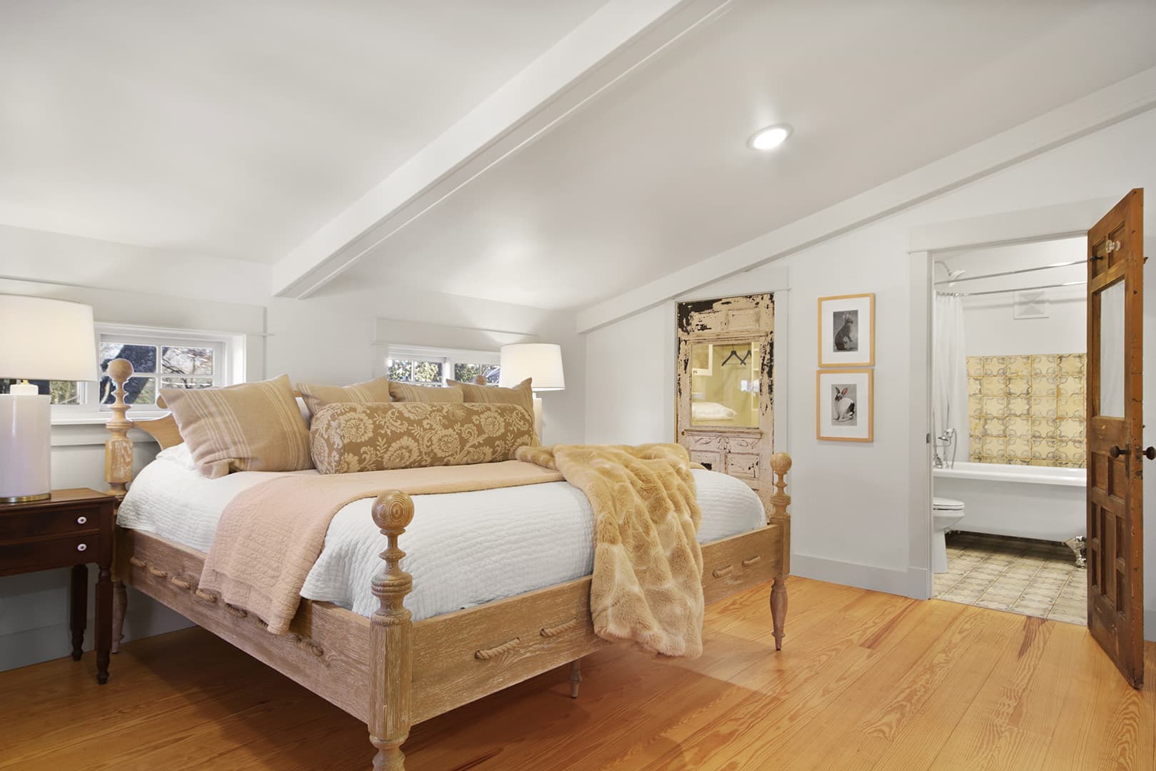 A serene attic bedroom with a light wood floor, a large bed with patterned bedding, and a glimpse into an adjoining bathroom.