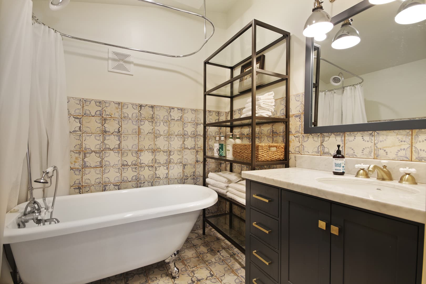 A charming bathroom with a clawfoot tub, patterned tiled walls, a dark vanity with gold hardware, and open metal shelving.