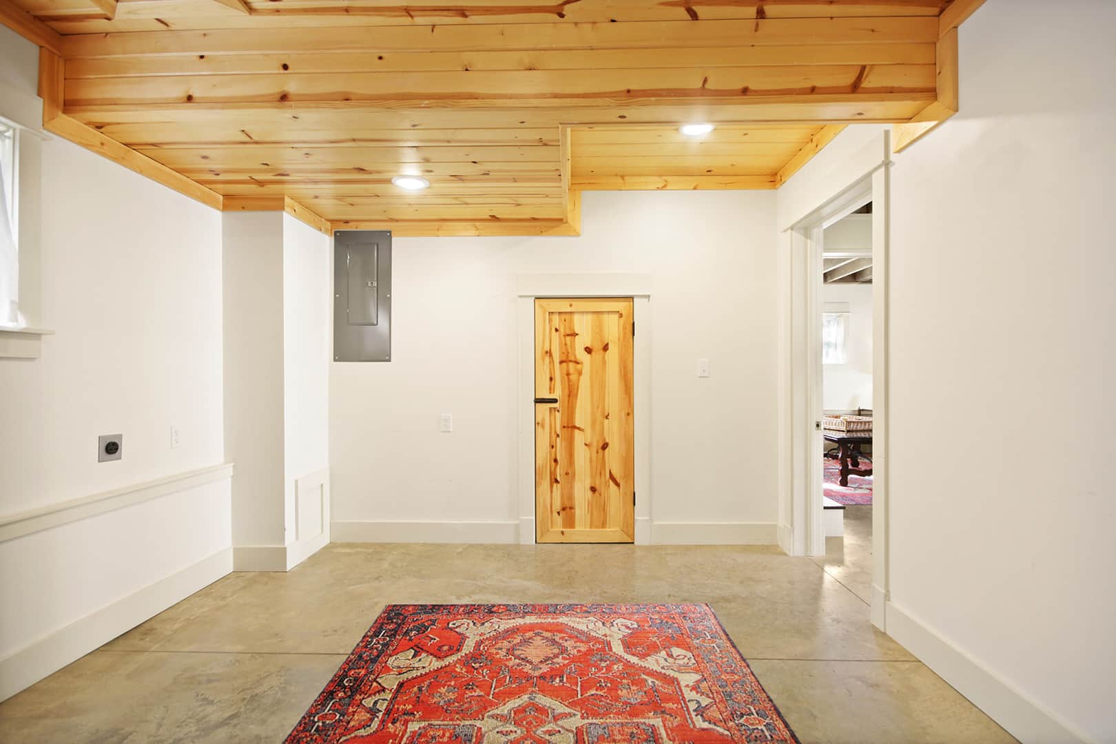 A spacious entryway with a light wood ceiling, a wooden door, and a vibrant red patterned rug on a light-colored floor.