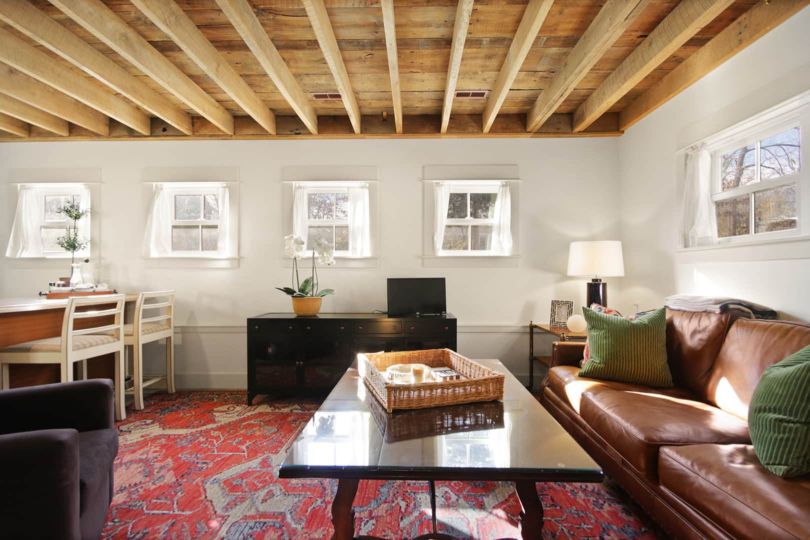 A rustic living space with exposed wooden ceiling beams, a brown leather sofa, a red oriental rug, and multiple windows.