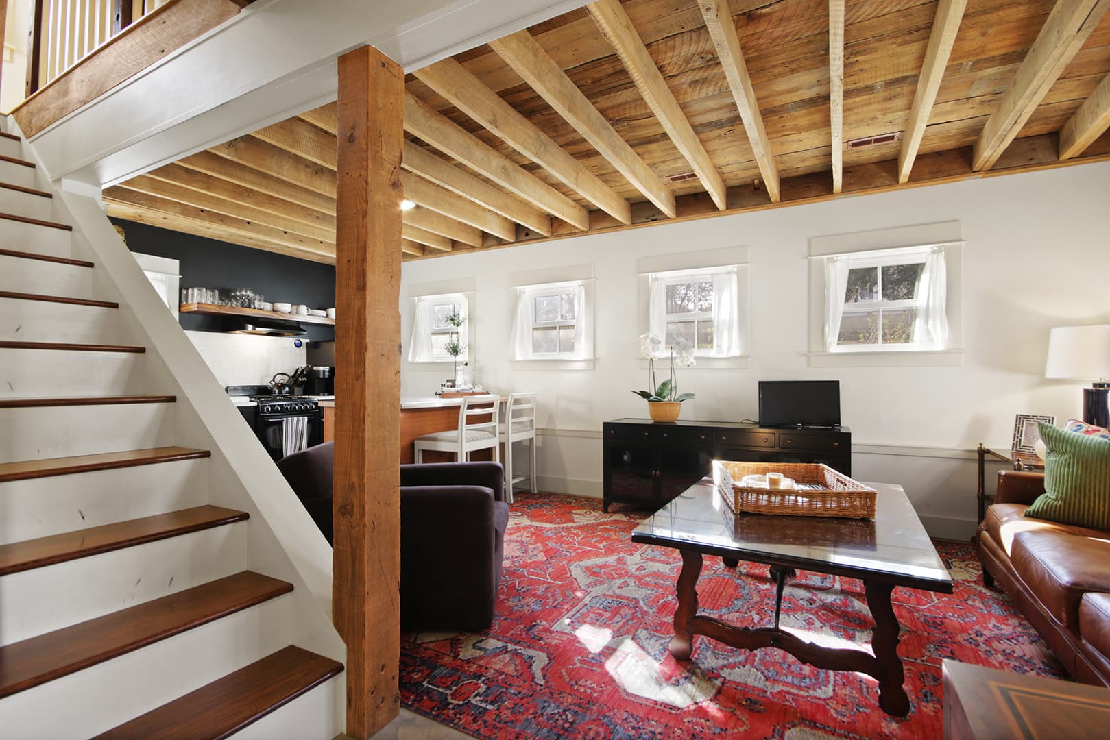 A cozy living area with exposed wooden ceiling joists, a brown leather couch, a vibrant red rug, and a staircase leading to another level.