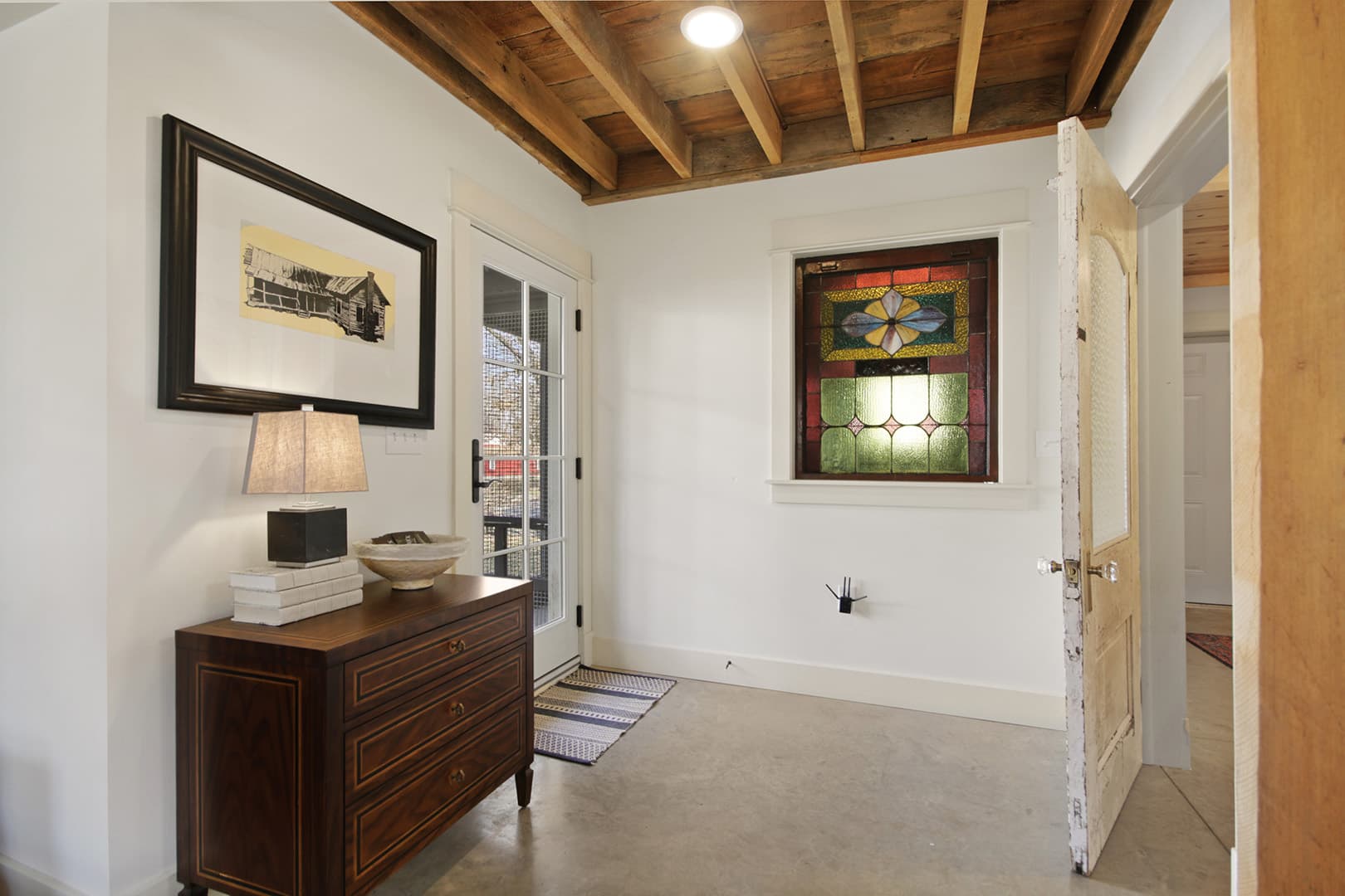 A rustic entry space with a wooden exposed-beam ceiling, cream walls, a dark wood dresser, and a vibrant stained-glass window.