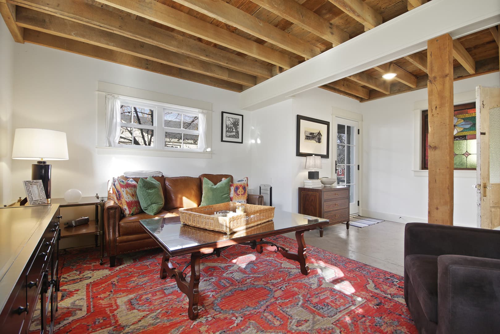 A stylish living room with exposed wooden ceiling joists, a leather sofa, a dark coffee table, and a vibrant red patterned rug.