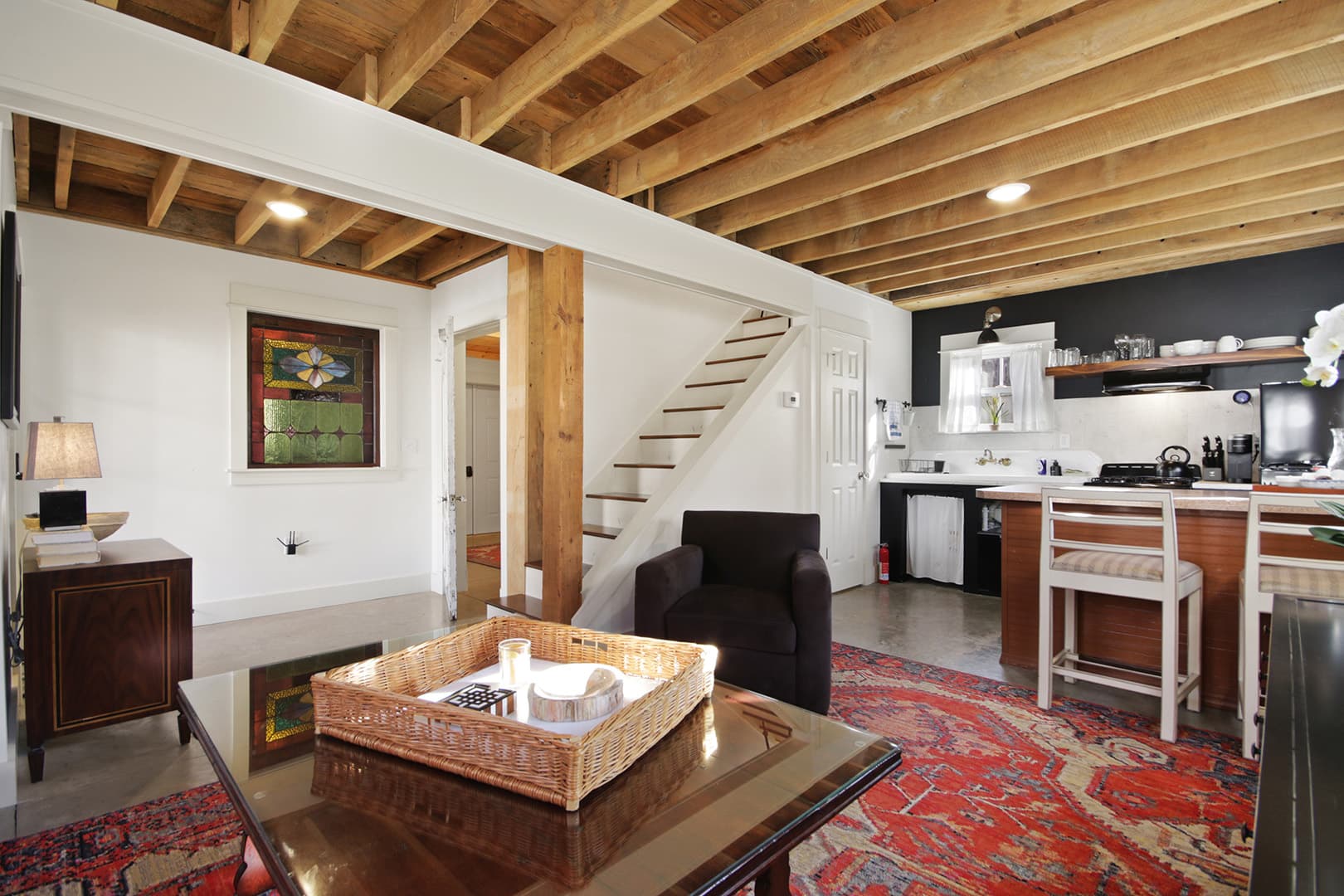 A spacious open-concept room with exposed wooden ceiling joists, a red patterned rug, and a staircase leading to a higher level.