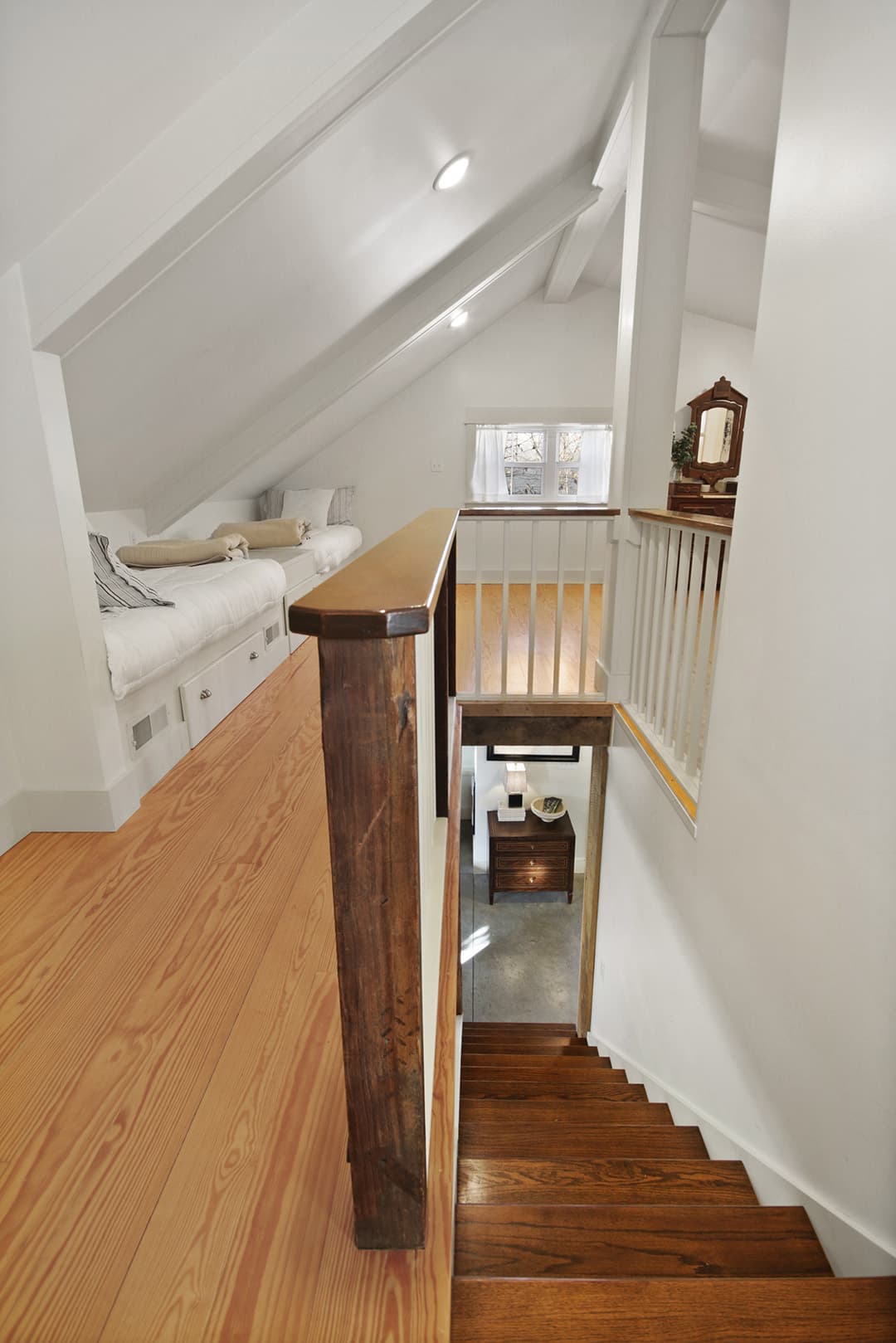 A warm and inviting loft bedroom with sloped ceilings, light wood floors, and a view down a wooden staircase.