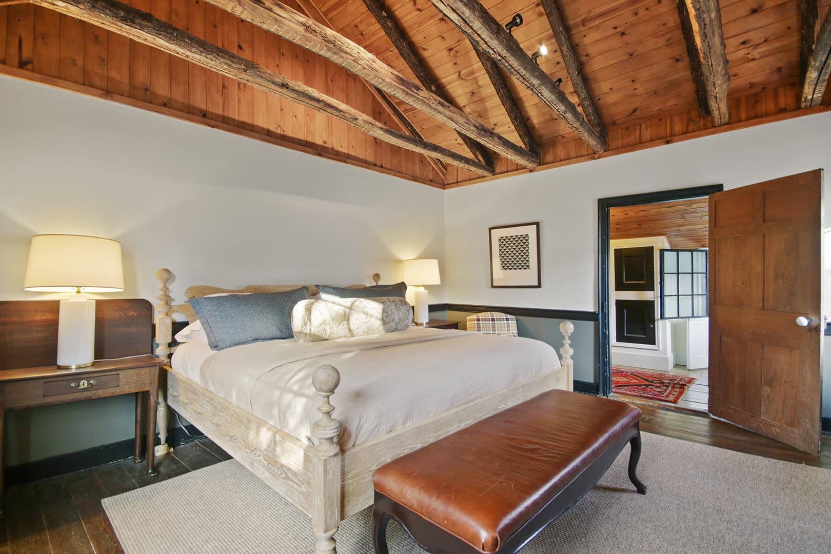 A stylish bedroom featuring a large bed with a light headboard, a wooden beamed ceiling, and a brown leather bench at the foot of the bed.