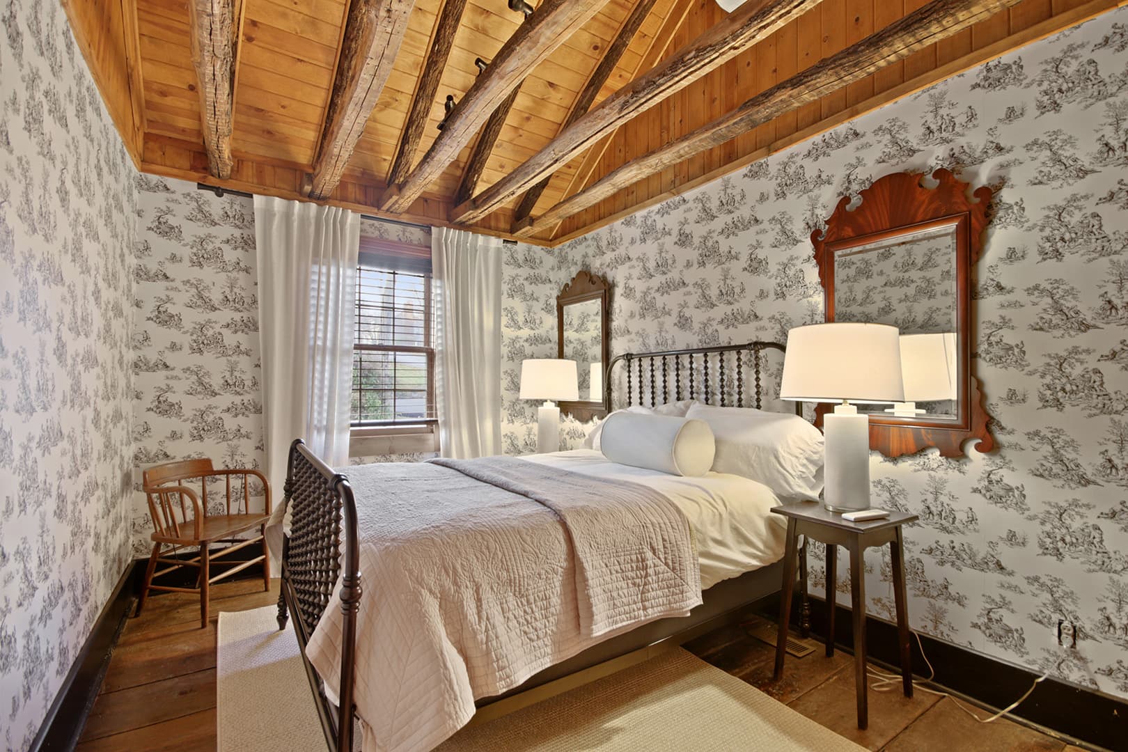 A charming bedroom featuring a bed with white and light pink bedding, a striking patterned wallpaper, and a rustic wooden beamed ceiling.