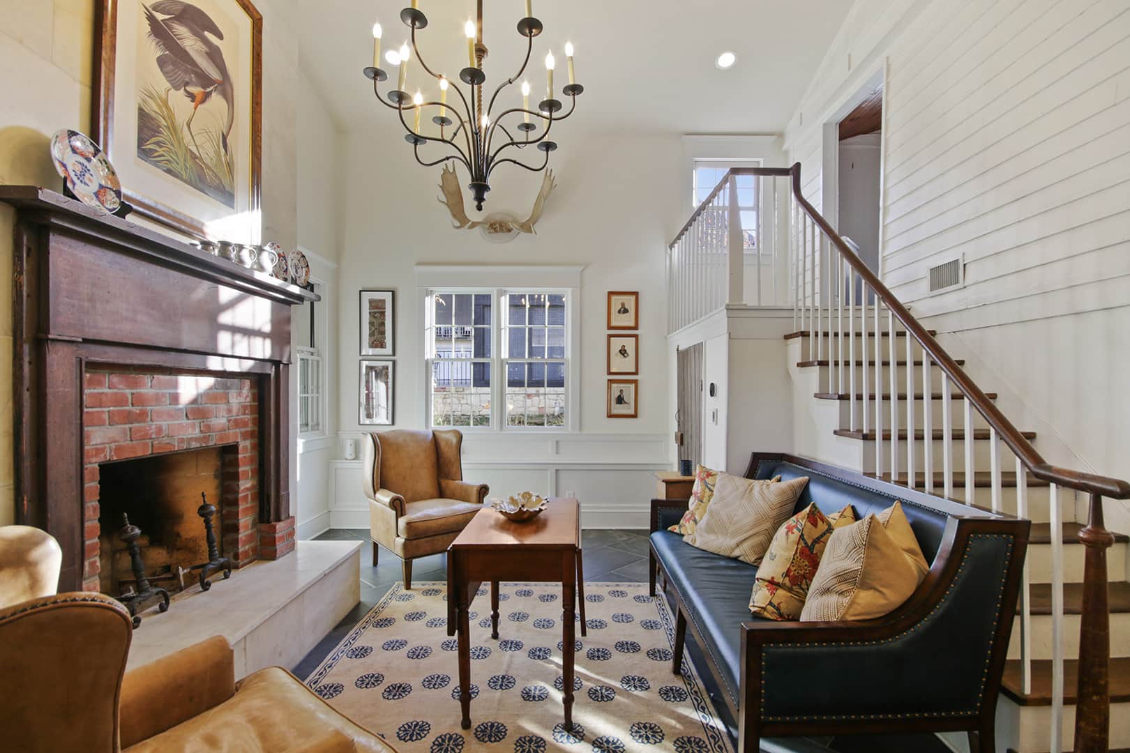 A classic living room with a fireplace, dark wood mantel, high ceilings, a grand chandelier, and a prominent staircase.