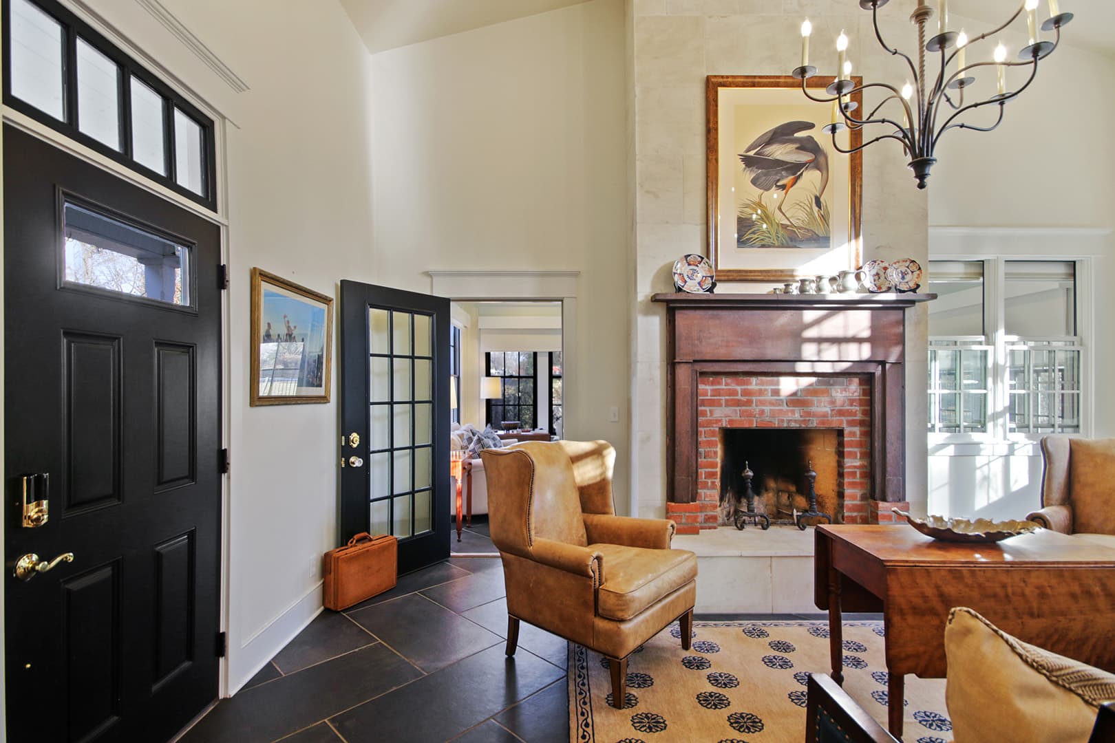 A stately living room with a black front door, a brick fireplace, leather armchairs, and a large chandelier illuminating the space.