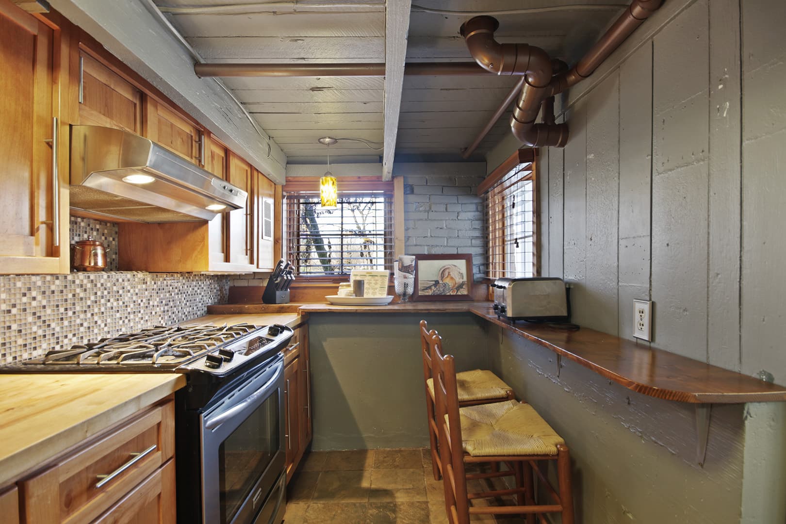 A galley-style kitchen with wood cabinets, a stove, a bar counter with stools, and exposed piping on the ceiling.