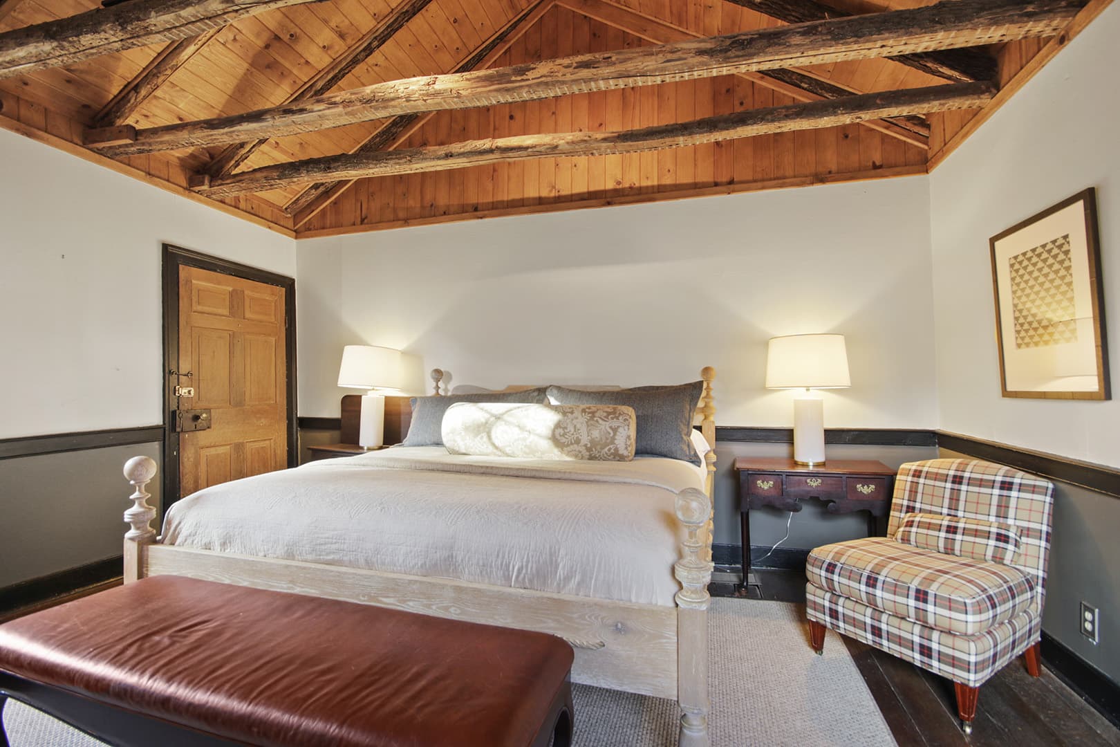 A luxurious bedroom featuring a king-sized bed, exposed wooden beam ceiling, dark wood accents, and a plaid armchair.