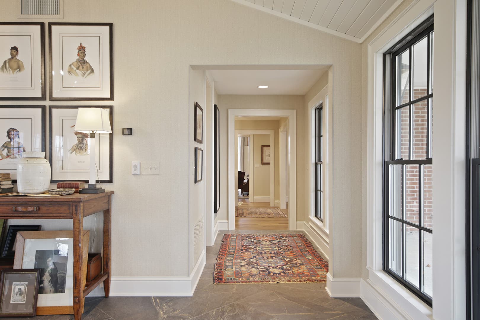 A bright hallway with a traditional aesthetic, featuring framed artwork, a console table, and a patterned rug leading to another room.