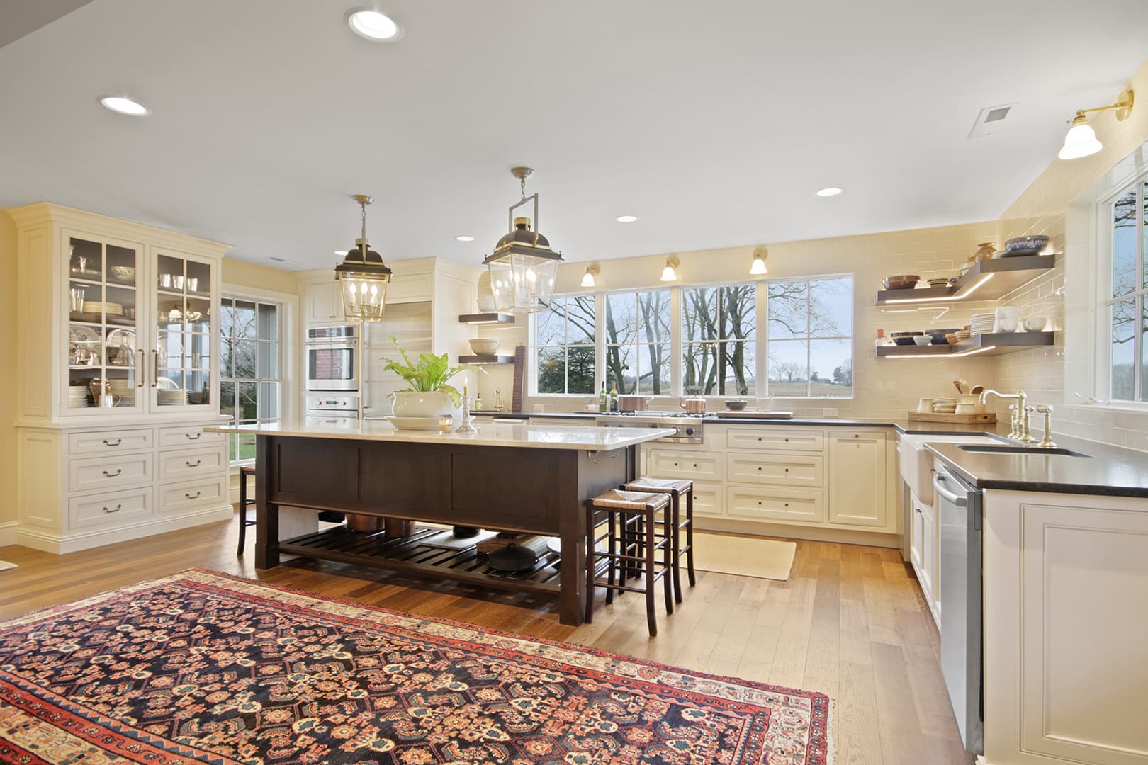 A spacious, bright open-concept kitchen with a large island, traditional cabinetry, modern lighting, and a decorative rug on hardwood floors.