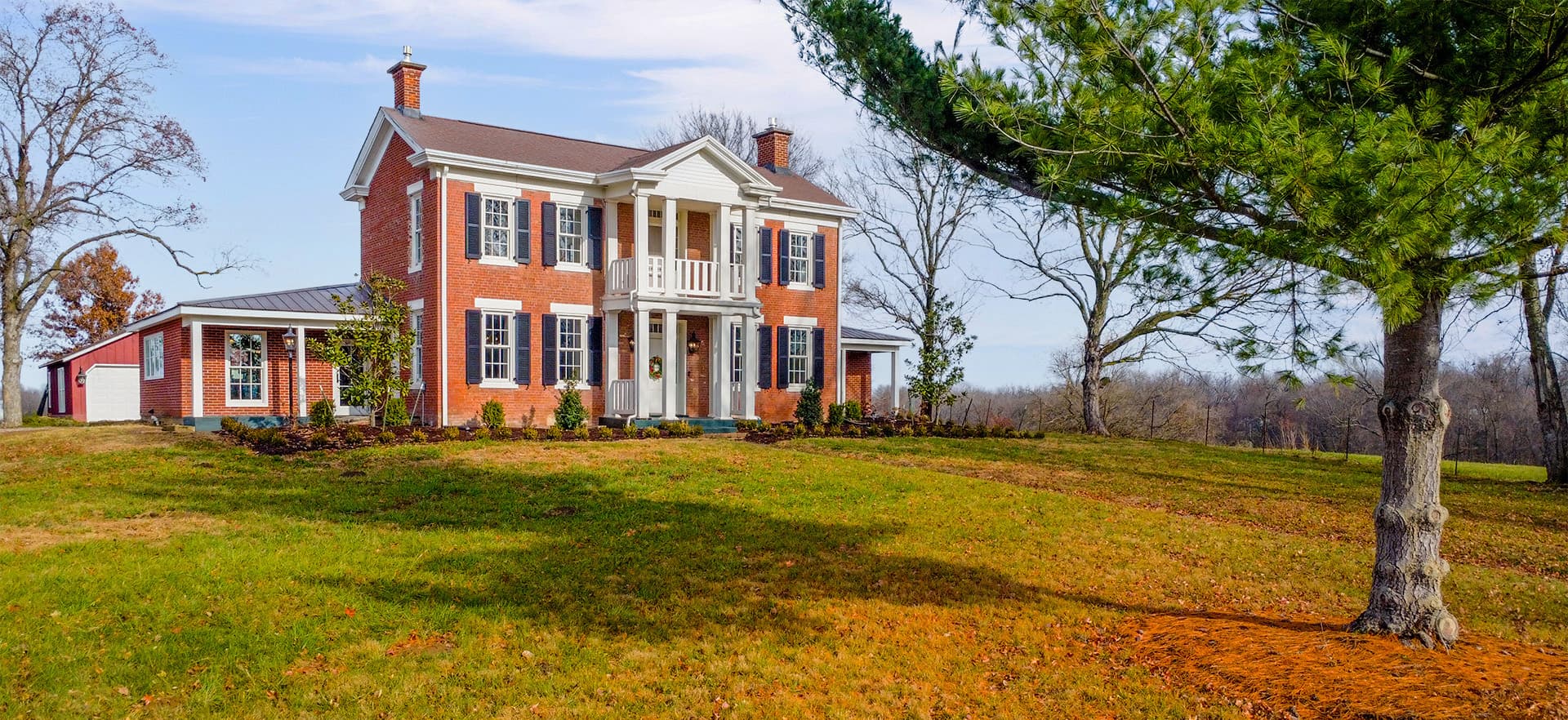 A two-story red brick house with white trim and a large front porch, surrounded by a grassy lawn and trees.